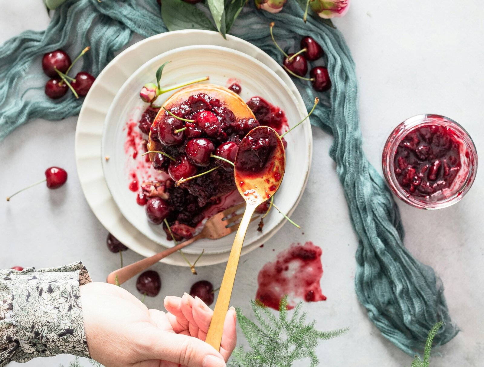 zdjęcie przedstawiające bezglutenowe pancakes z wiśniami i rękę kobiety, która dokłada więcej konfitury