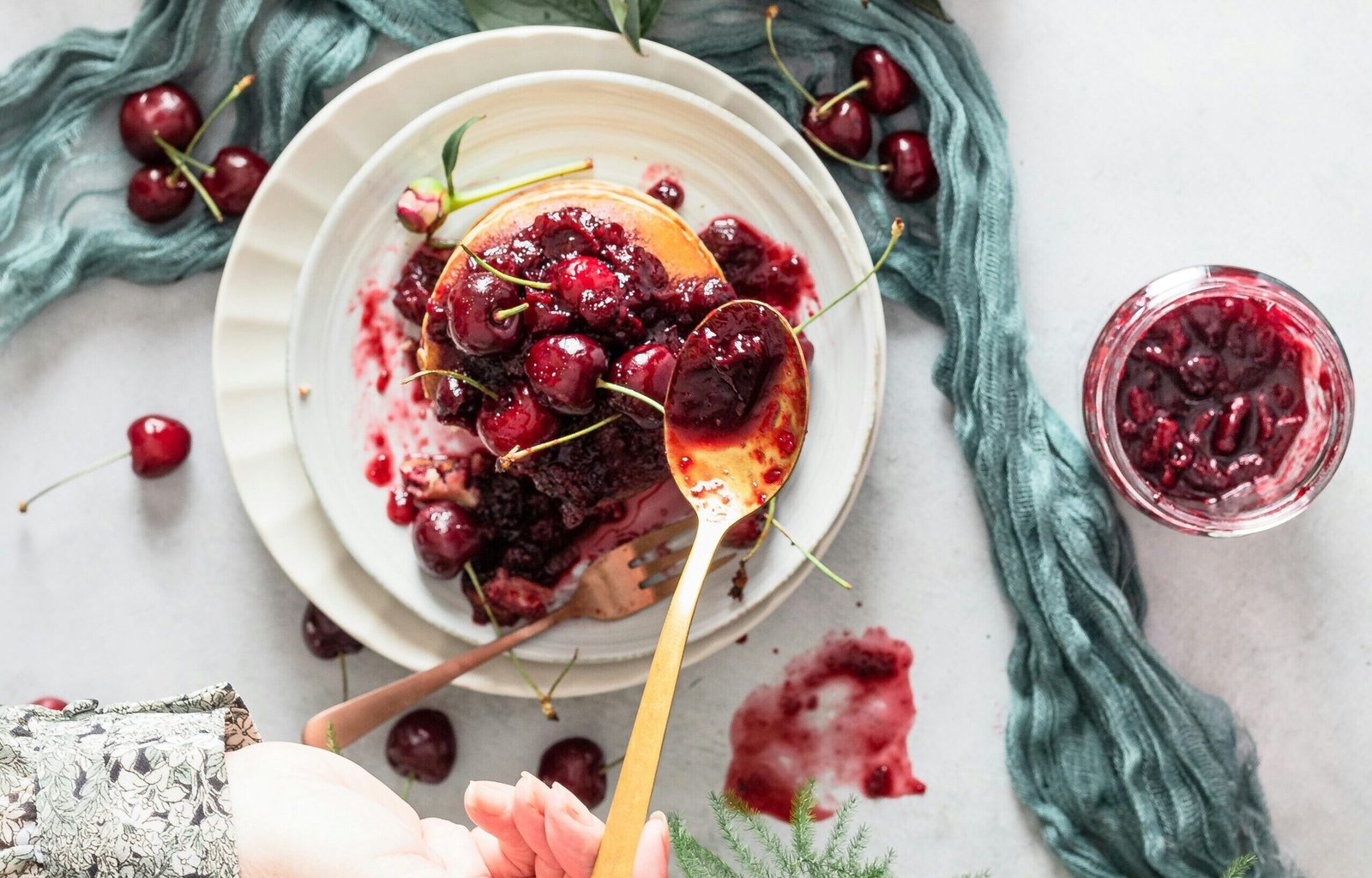 zdjęcie przedstawiające bezglutenowe pancakes z wiśniami i rękę kobiety, która dokłada więcej konfitury