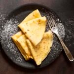 zdjęcie naleśników bez glutenu ułożonych na sobie na talerzu, posypanych cukrem pudrem i z widelcem obok