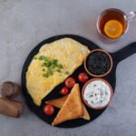 zdjęcie omletu z serka wiejskiego podanego na czarnej podstawce w towarzystwie szczypiorku, tostów, pomidorków, białego serka, kawioru i herbaty z cytryną