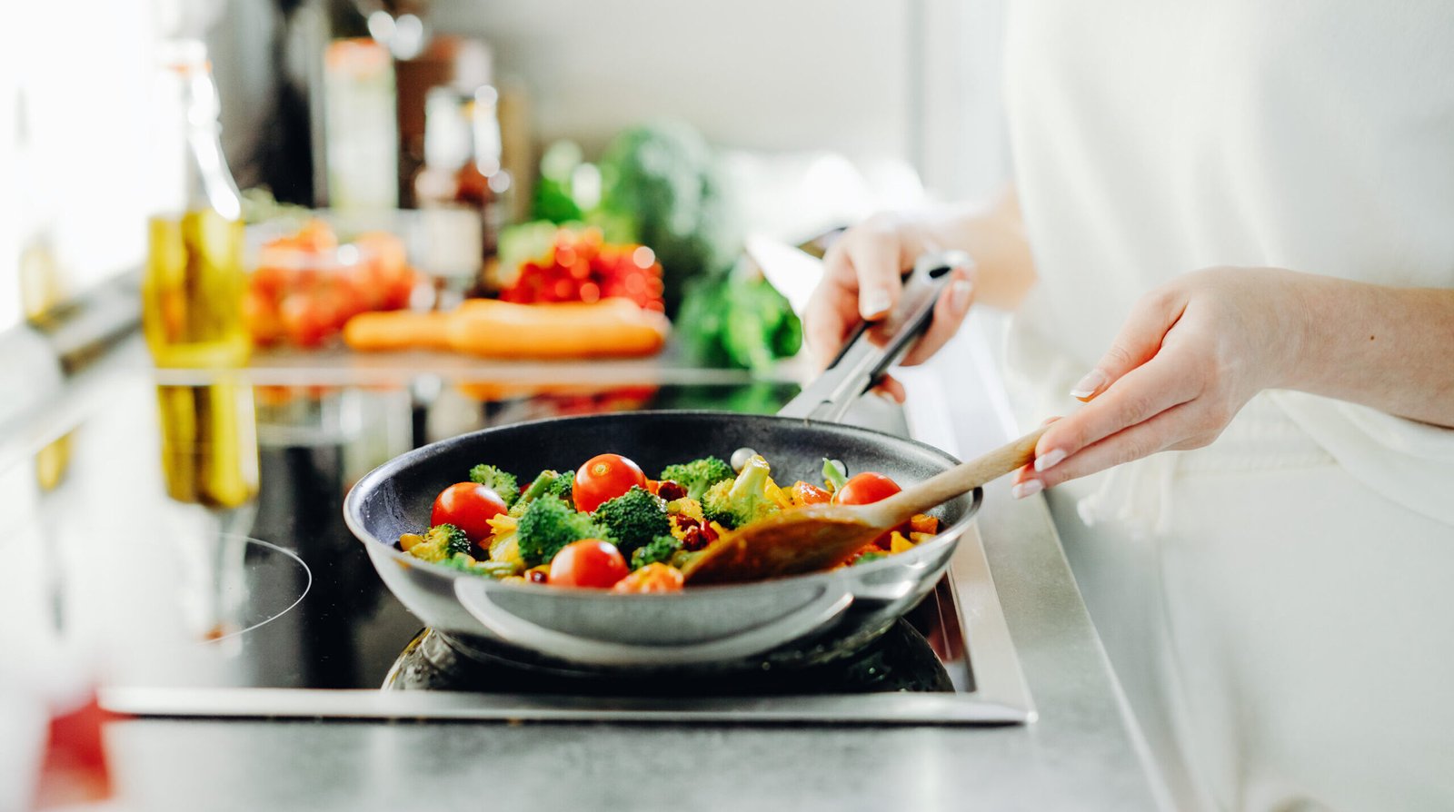 zdjęcie przestawiające ręce kobiety, trzymającej patelnię i smażącej warzywa na kuchence indukcyjnej, w tle przybory kuchenne i warzywa