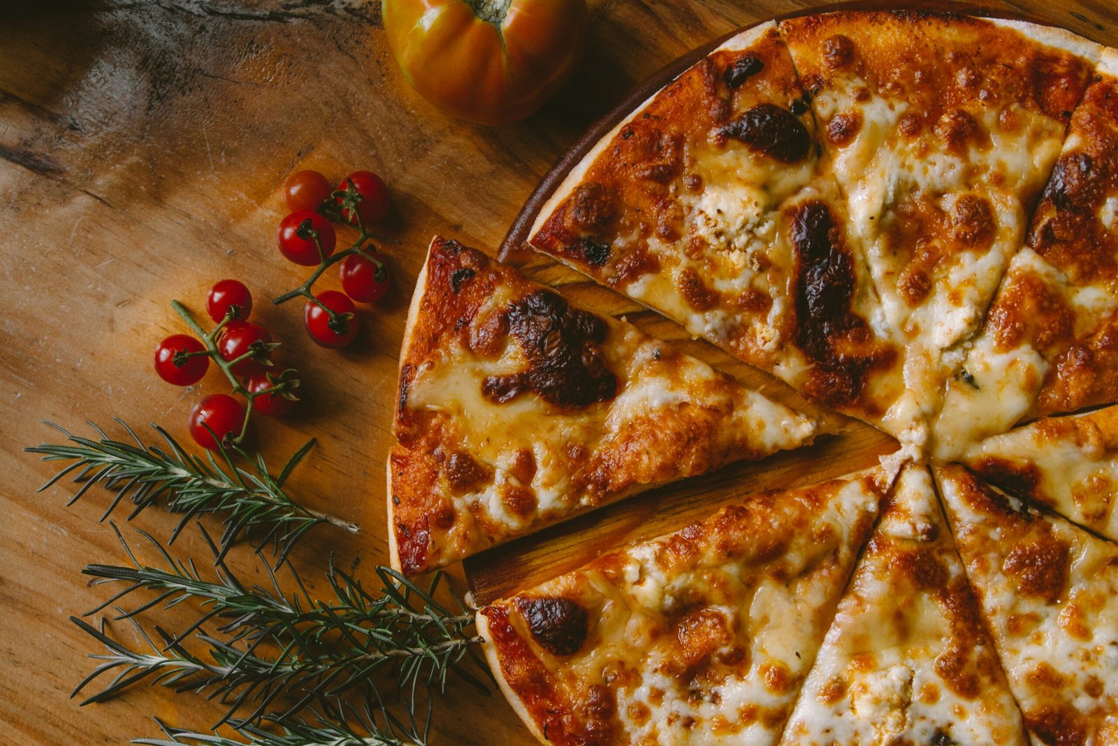 zdjęcie pokrojonej pizzy na desce na drewnianym stole, na którym leży tez rozmaryn, pomidor i pomidorki koktailowe