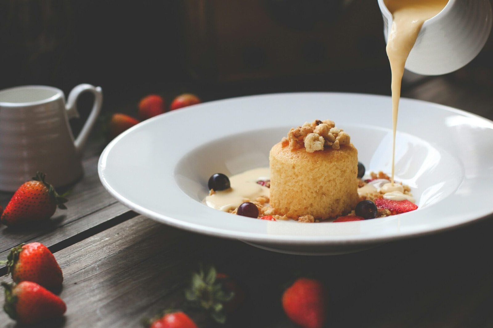 zdjęcie sernika na białym głębokim talerzu w towarzystwie truskawek i winogron oraz orzechów, po prawej widać biały dzbanek z którego wylewa się kremowy sos na talerz z sernikiem. W tle widać ciemny stół, na nim truskawki i biały dzbanuszek