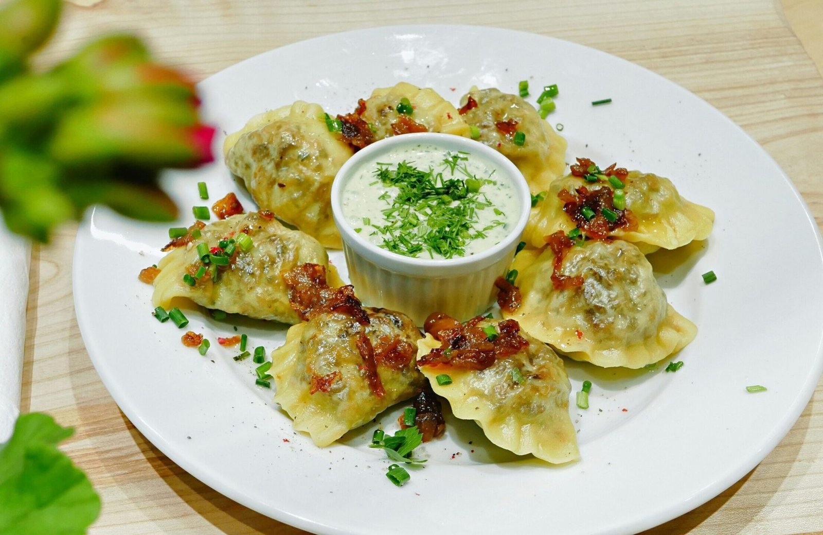 zdjęcie bezglutenowych pierogów na białym talerzu, posypanych cebulką zasmażoną i szczypiorkiem z białym sosem na środku