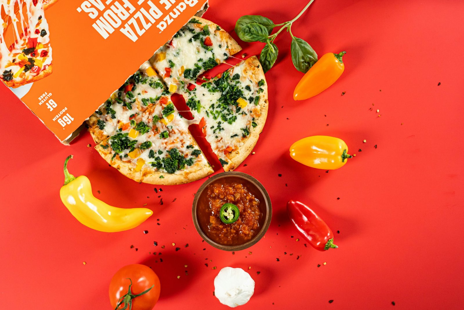 Bezglutenowa pizza podzielona na cztery części , wokół niej znajdują się różne warzywa, sos i przyprawy, wszystko znajduje się na czerwonym tle