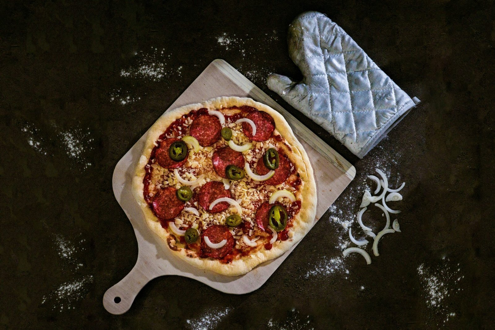 zdjęcie bezglutenowej pizzy z salami jalapino i cebulą ułożonej na drewnianej desce do krojenia obok rękawicy kuchennej