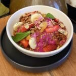 Miseczka smoothie bowl w intensywnym różowym kolorze, udekorowana truskawkami, bananem, granolą bezglutenową, płatkami migdałów, nasionami chia, listkami mięty i jadalnymi kwiatami