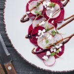 Carpaccio z buraka bez glutenu, podane z rzodkiewką, serem feta, kiełkami i sosem balsamicznym na białym talerzu.