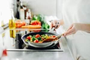 zdjęcie przestawiające ręce kobiety, trzymającej patelnię i smażącej warzywa na kuchence indukcyjnej, w tle przybory kuchenne i warzywa