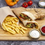 Domowy kebab w bezglutenowej tortilli, pokrojony na połówki i zawinięty w papier, podany z porcją frytek oraz sosem ketchupowym i czosnkowym na drewnianej desce