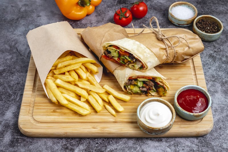 Domowy kebab w bezglutenowej tortilli, pokrojony na połówki i zawinięty w papier, podany z porcją frytek oraz sosem ketchupowym i czosnkowym na drewnianej desce