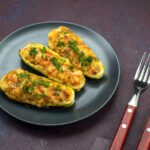 Trzy połówki grillowanej cukinii nadziewane mięsem i warzywami, zapieczone z serem i posypane świeżymi ziołami, podane na ciemnym talerzu obok widelca i noża.