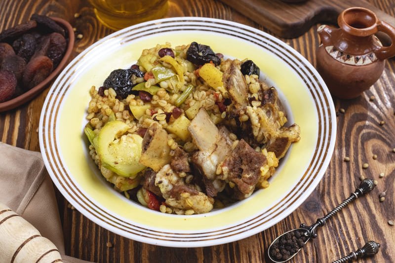 Miska gulaszu z kawałkami mięsa, warzywami i ziarnami, podana na drewnianym stole obok suszonych owoców i tradycyjnych naczyń