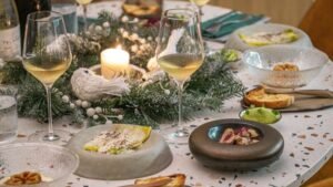 Wigilijny stół z elegancką zastawą, zapalona świeca w stroiku z gałązek świerku, kieliszki z białym winem oraz półmiski z potrawami i pieczywem ustawione na jasnym blacie stołu.