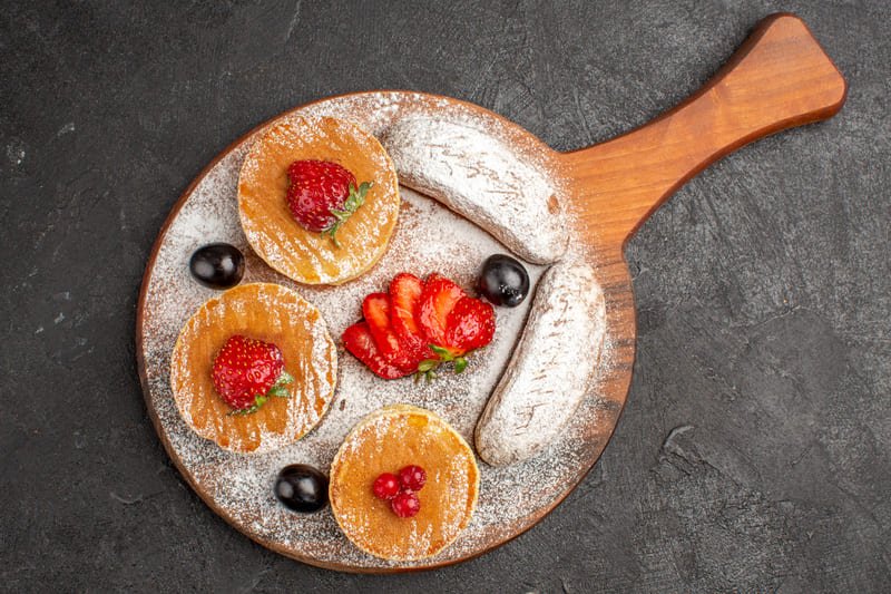 Bezglutenowe placuszki twarogowe podane na drewnianej desce, oprószone cukrem pudrem i udekorowane truskawkami, winogronami oraz czerwonymi owocami.