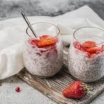 Dwa szklane pucharki puddingu chia na mleku z kawałkami truskawek, ustawione na drewnianej desce, z dodatkowymi owocami obok.