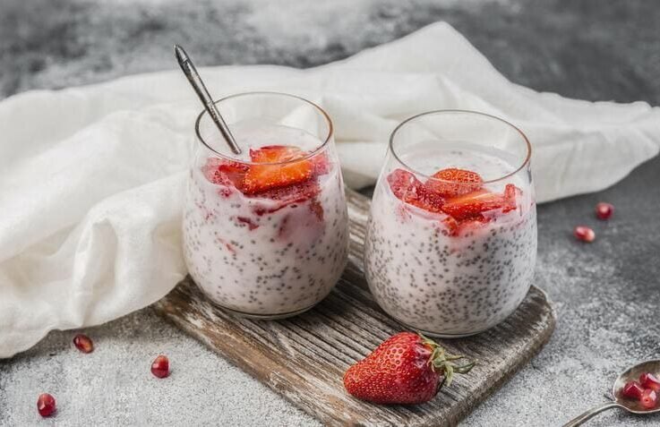 Dwa szklane pucharki puddingu chia na mleku z kawałkami truskawek, ustawione na drewnianej desce, z dodatkowymi owocami obok.