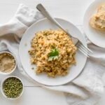 Talerz z bezglutenowym risotto z warzywami, podany na białym stole z kawałkami pieczywa bezglutenowego i miseczkami z przyprawami oraz zieloną soczewicą.