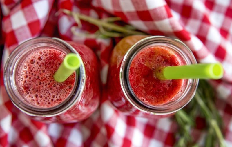 Dwie szklane butelki wypełnione czerwonym koktajlem owocowym z zielonymi słomkami, ustawione na czerwono-białej kraciastej tkaninie.