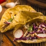 Bezglutenowe tortille kukurydziane wypełnione warzywami, czerwoną kapustą, rzodkiewką i ryżem, podane na drewnianym stole z papryczką chili i czosnkiem w tle.