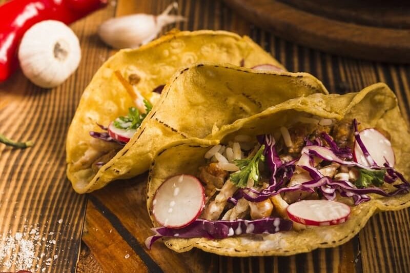 Bezglutenowe tortille kukurydziane wypełnione warzywami, czerwoną kapustą, rzodkiewką i ryżem, podane na drewnianym stole z papryczką chili i czosnkiem w tle.