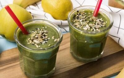 Dwa szklane kubki z zielonym koktajlem warzywno-owocowym posypanym mieszanką nasion, ustawione na drewnianej desce.