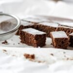 zdjęcie brownie bez glutenu pokrojonego na kawałki i posypanego cukrem pudrem, ułożonego na białej powierzchni obok metalowego sitka