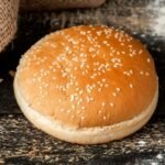 bezglutenowa bułka do burgerów posypana sezamem, upieczona na złoto, podana na ciemnym, rustykalnym blacie.