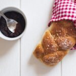 zdjęcie chałki owiniętej w ściereczkę kratkę od góry, obok w kokilce stoi dżem z łyżeczką w środku