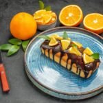 Bezglutenowe ciasto pomarańczowe polane czekoladową polewą, udekorowane kawałkami świeżej pomarańczy, podane na niebieskim talerzu, obok całe i przekrojone pomarańcze na ciemnym tle.