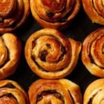 Bezglutenowe bułeczki cynamonowe w formie złocistych ślimaczków z nadzieniem cynamonowym i rodzynkami, upieczone na blasze