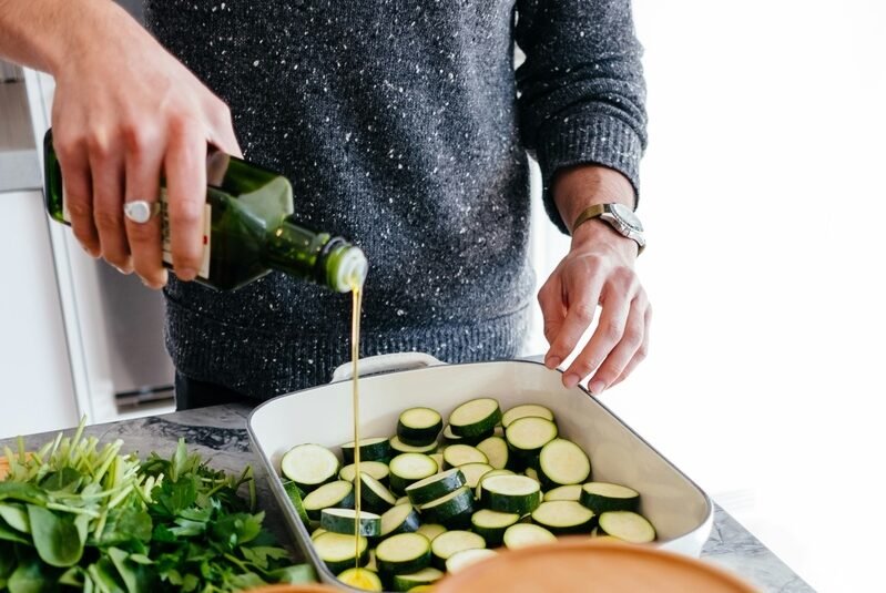 zdjęcie przedstawiające część mężczyzny który polewa oliwą warzywa w naczyniu do zapiekania w kuchni