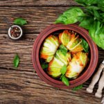 zdjęcie gołąbków bez glutenu w sosie pomidorowym w glinianym naczyniu postawionym na drewnianym stole obok sztućców, słoiczka przypraw oraz bazylii