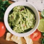 zdjęcie guacamole w białej miseczce ustawionej na drewnianej desce w towarzystwie limonki, pomidorków oraz pietruszki