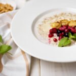 Bezglutenowa jaglanka na mleku z karmelizowanym bananem, malinami, porzeczkami i listkiem mięty, podana na białym talerzu.