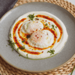 zdjęcie jajek po turecku bez glutenu, na jogurcie, z masłem paprykowym i koperkiem, podane na szarym talerzu, ustawionym na plecionej podkładce na stole