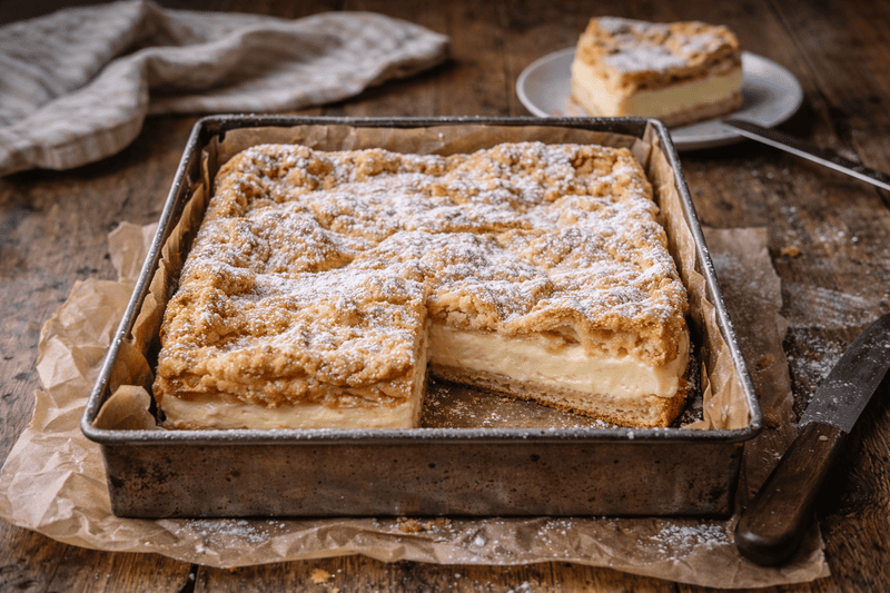Bezglutenowa karpatka upieczona w kwadratowej blaszce, z puszystym ciastem parzonym i kremem budyniowym, podana w domowym otoczeniu.