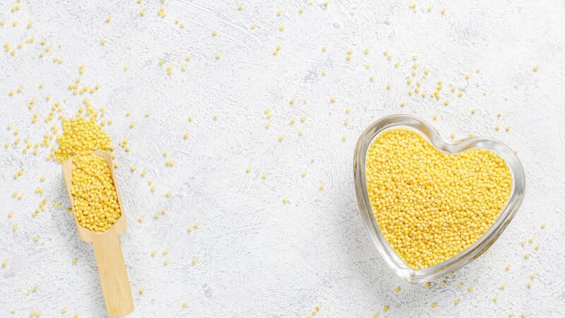 Sucha kasza jaglana w drewnianej misce i łopatce, rozsypana na jasnym, drewnianym blacie.