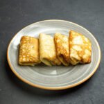 Cztery bezglutenowe krokiety złożone z naleśników, ułożone obok siebie na szarym talerzu z beżowym rantem