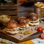 Miniburgery bez glutenu na drewnianej desce, z bułkami posypanymi sezamem, kremowym sosem i warzywami, idealne na sylwestrową imprezę.