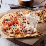 bezglutenowa cienka pizza z dodatkiem ciągnącego się sera, sosu pomidorowego i cebuli ułożona na papierze do pieczenia na drewnianym, udekorowanym stole