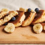 zdjęcie bezglutenowych placków bananowych z z płatkami owsianymi podanych na drewnianej podstawce z kawałkami bananów, borówkami oraz bezglutenowym sosem czekoladowym