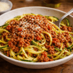 Bezglutenowe spaghetti bolognese z makaronem z cukinii, podane na białym talerzu z sosem pomidorowo-mięsnym i startym parmezanem.