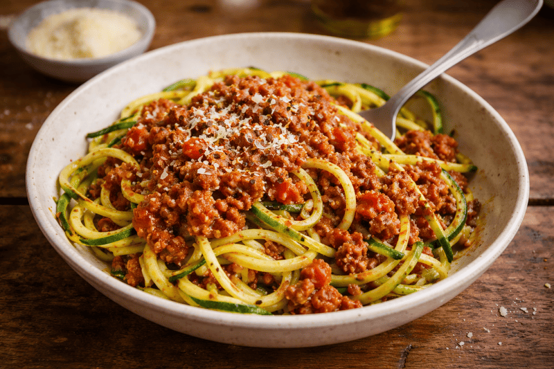 Bezglutenowe spaghetti bolognese z makaronem z cukinii, podane na białym talerzu z sosem pomidorowo-mięsnym i startym parmezanem.