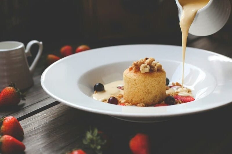 zdjęcie sernika na białym głębokim talerzu w towarzystwie truskawek i winogron oraz orzechów, po prawej widać biały dzbanek z którego wylewa się kremowy sos na talerz z sernikiem. W tle widać ciemny stół, na nim truskawki i biały dzbanuszek