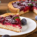 Kruche ciasto bezglutenowe ze śliwkami, posypane cukrem pudrem, podane na białym talerzyku z widoczną całą tartą w tle.