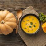 zdjęcie naturalnie bezglutenowej zupy dyniowej podanej w misce z pestkami dyni, natką pietruszki i kleksem mleczka kokosowego. Na stole obok zupy leży dynia i drewniana łyżka