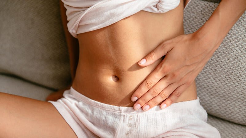 Osoba siedząca na kanapie, trzymająca dłoń na brzuchu – ilustracja wzdęć i dyskomfortu brzucha, które mogą towarzyszyć celiakii i reakcji organizmu na gluten.