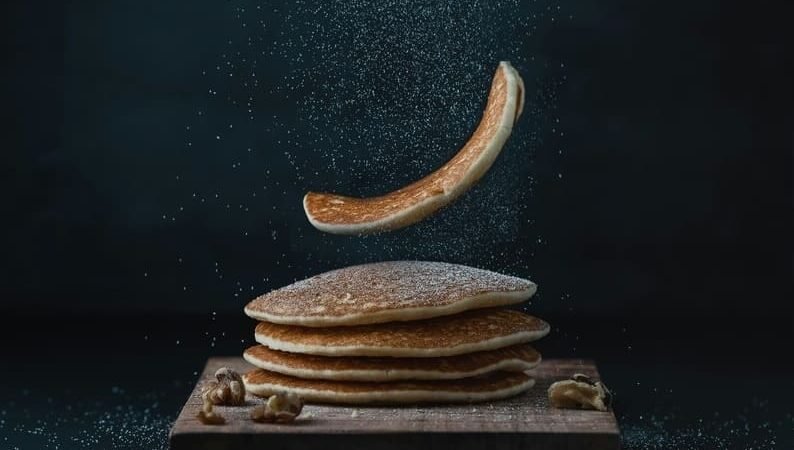 Naleśniki z mąki kukurydzianej bez glutenu ułożone na drewnianej desce, oprószone cukrem pudrem, z unoszącym się w powietrzu naleśnikiem – inspiracja do bezglutenowych dań z mąki kukurydzianej.