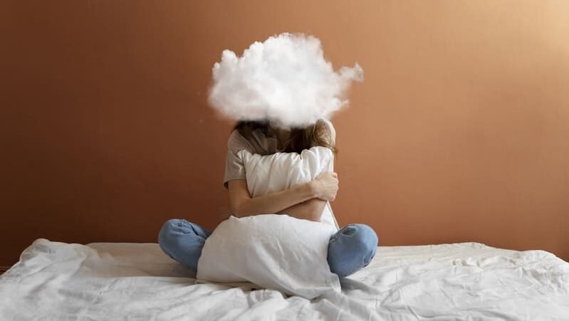 Osoba siedząca na łóżku, obejmująca poduszkę, z symboliczną chmurą nad głową – ilustracja objawów zatrucia glutenem, takich jak złe samopoczucie, zmęczenie i reakcja organizmu po spożyciu glutenu.